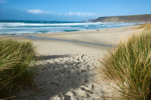 This landscape photograph depicts Sennen Cove beach in Cornwall, United Kingdom, as seen from the sandy dunes that frame the approach to the coast. The image was taken in the early afternoon during the autumn season, with bright sunlight casting clear shadows over the beach and grass-tufted dunes. The wide expanse of sand leads to the rolling waves of the blue sea, and the distant headlands typical of the rugged Cornish coast are visible on the horizon. The natural setting highlights the scenic beauty of Sennen Cove and its coastal surroundings, offering a clear view of the relationship between the beach, the Atlantic Ocean, and the characteristic landscape of Cornwall in the United Kingdom.
